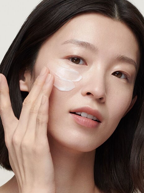 Woman applying white moisturizer to her cheekbone against grey background