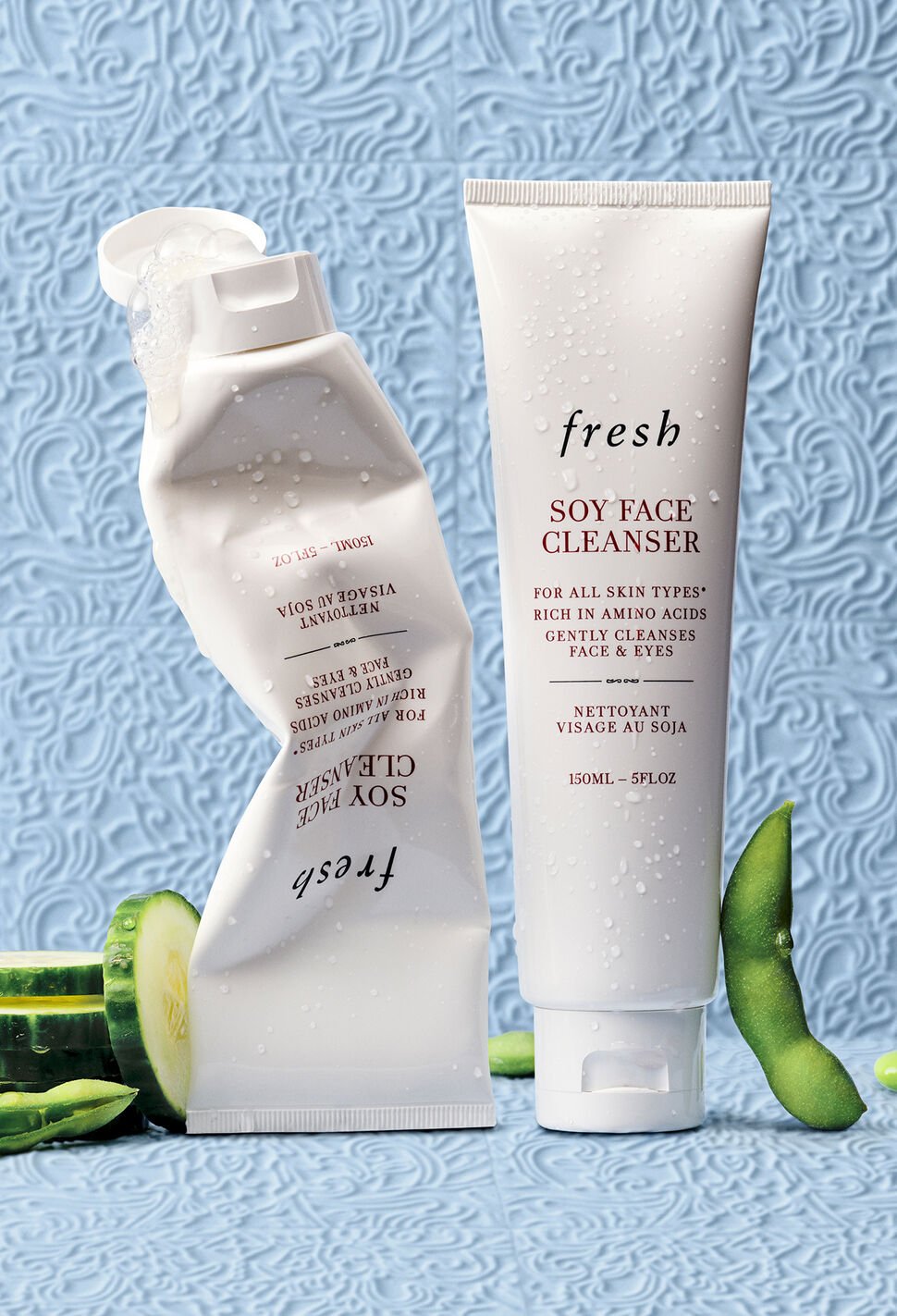 Crumpled and full Soy Face Cleanser tubes with sliced cucumber and soy bean on blue background